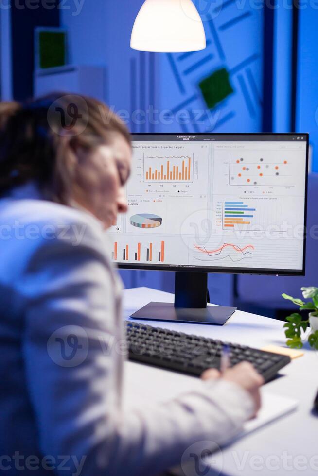 Tired entrepreneur trying to finish business project during internet videocall in front of computer, taking notes on notebook. Woman employee sitting at desk in workplace working at marketing ideas photo
