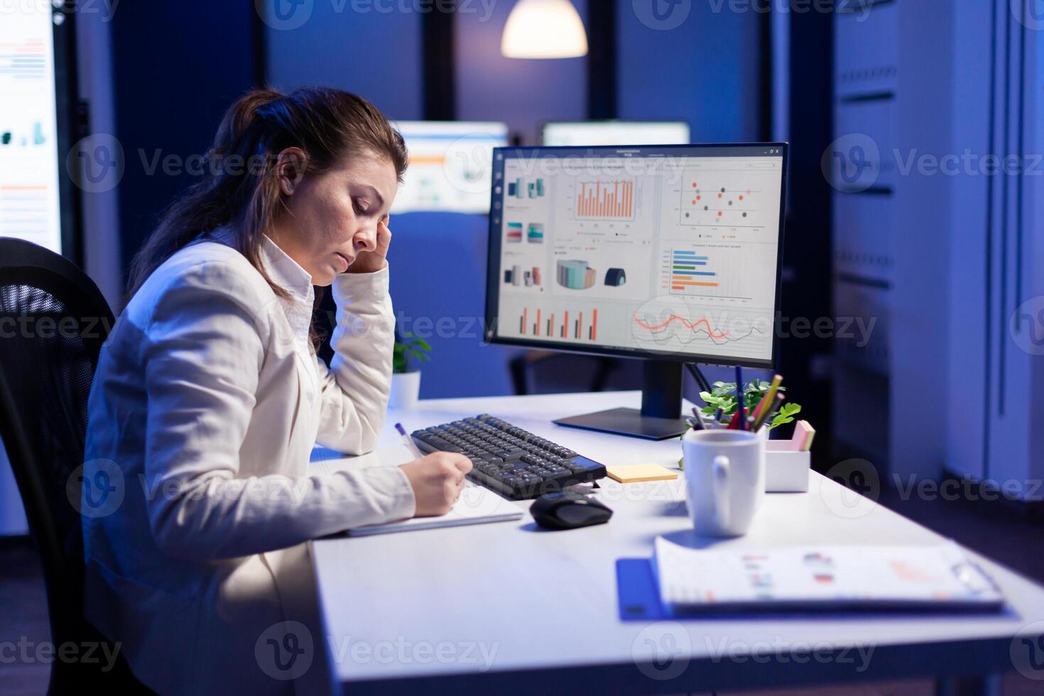 Back shot of overwhelmed woman working at night in front of computer, writing notes on notebook annual reports, checking financial deadline. Focused manager using technology network wireless photo