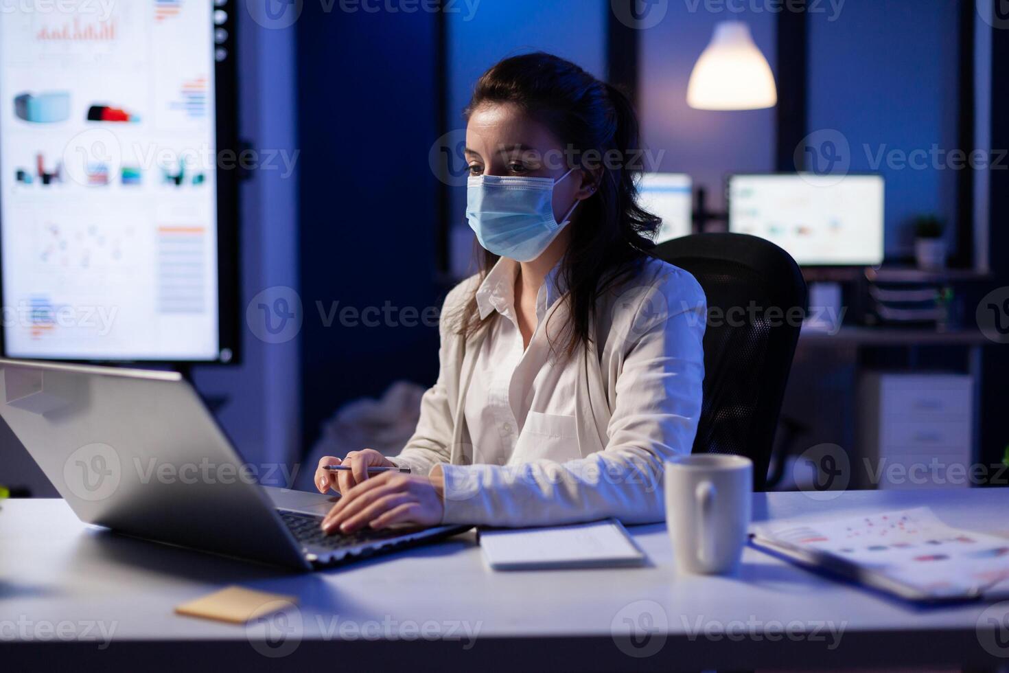 Manager woman with face mask working overtime in new normal business office looking at team project on professional laptop. Analysing financial documents sitting at desk during global pandemic photo