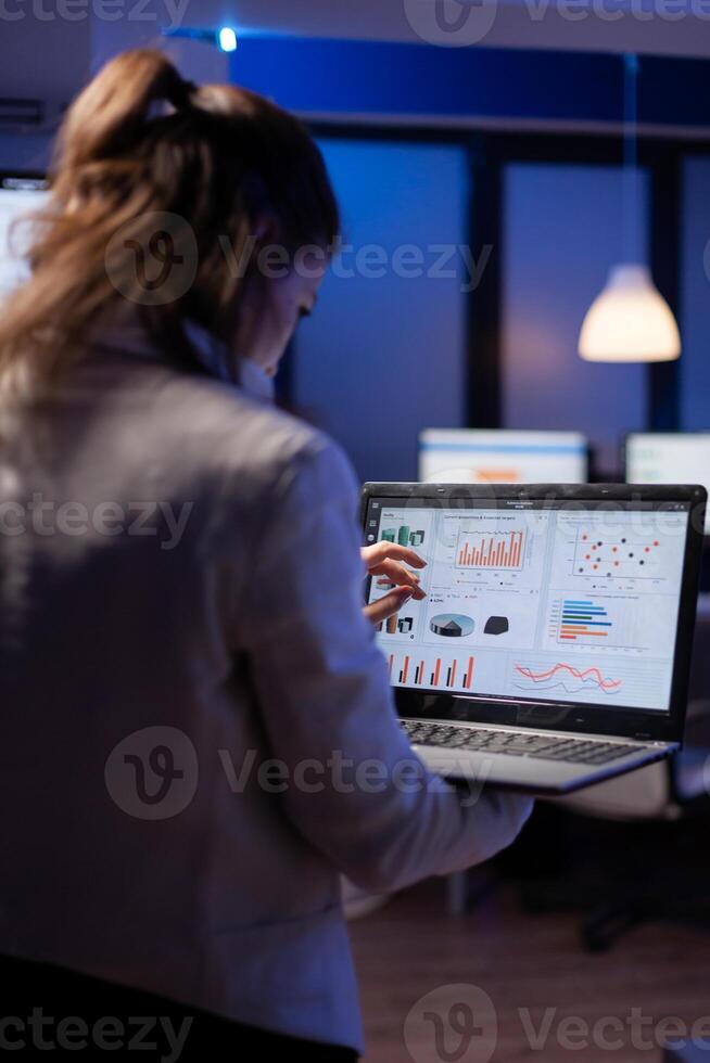 Back view of manager woman analysing financial statistics holding laptop standing in start-up business office late at night overworking. Tired focused employee using modern technology network wireless photo
