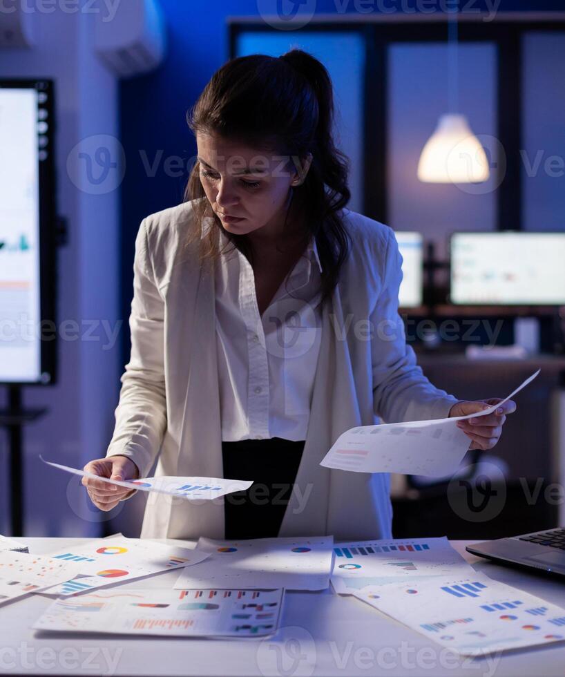 Stressed manager woman working with financial documents checking graphs, holding papers late at night. Business woman standing in start-up office doing overtime work to respect deadline. photo