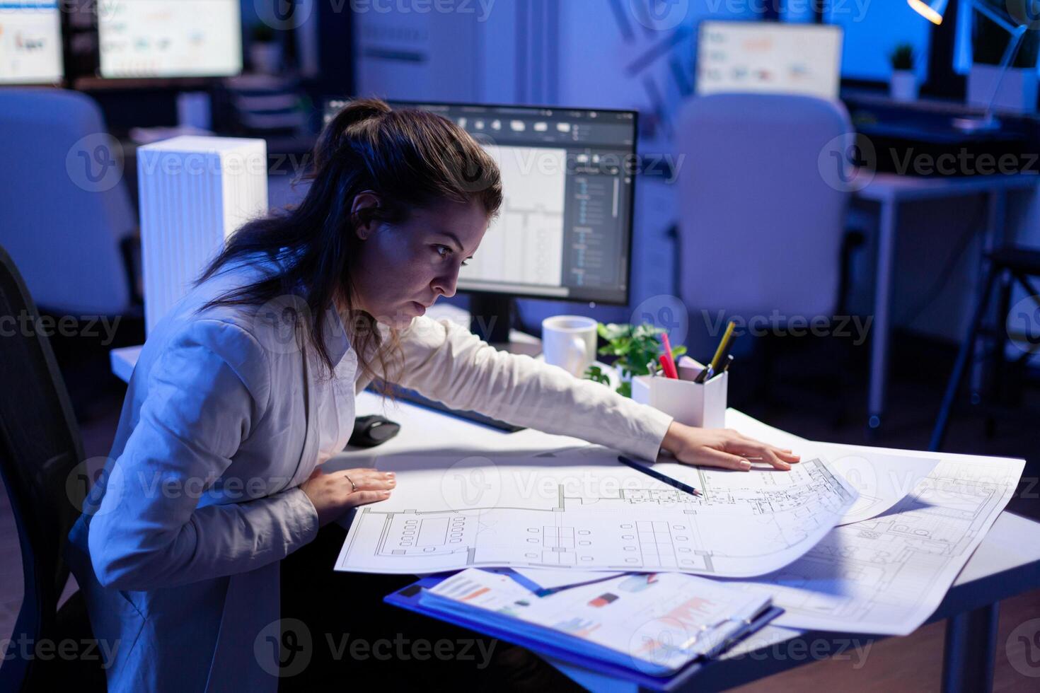 Overworked woman architect checking and matching blueprints sitting at office desk in front of computer. Perfectionist designer using arhitecture blueprints of buildings creating industrial prototype photo