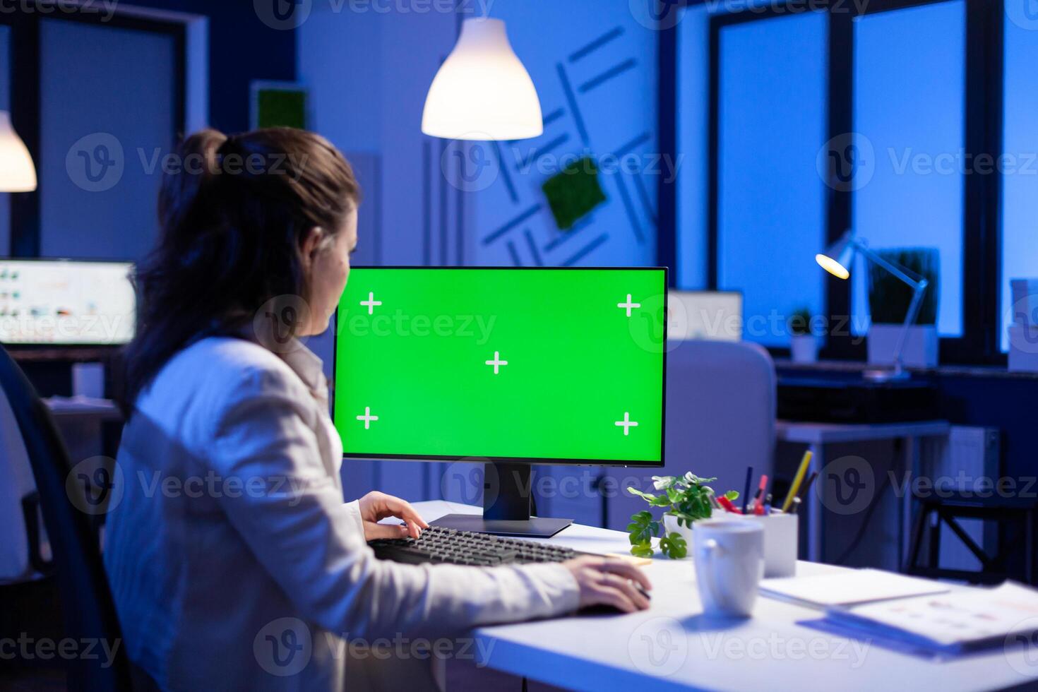 Entrepreneur working in front of green screen display sitting at desk during videocall meeting in business office. Businesswoman watching desktop monitor with green mockup, chroma key, working overtime. photo