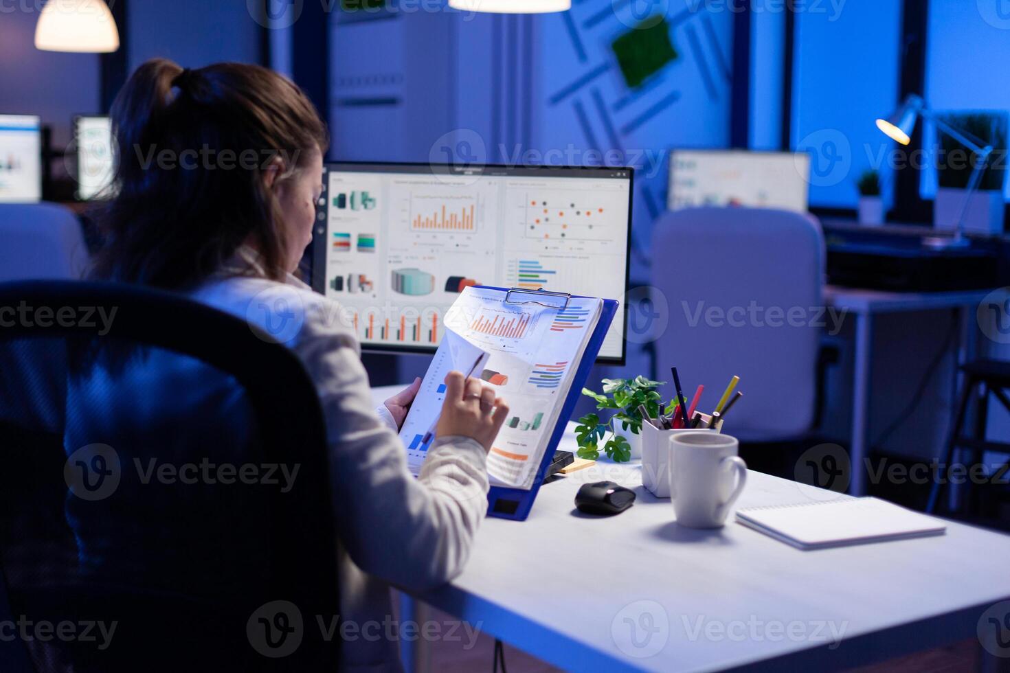 Freelancer woman comparing graphics from clipboard with grafics from computer at business office. Tired focused employee using modern technology network wireless working on project solution photo