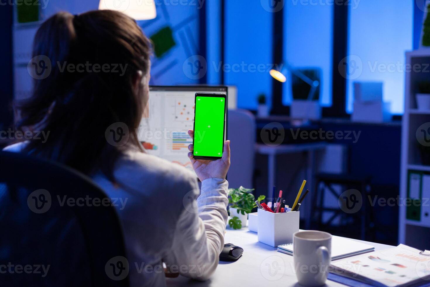 Entrepreneur looking at smartphone with green screen sitting at office desk overworking. Manager project watching desktop monitor display with mockup chroma key during videocall meeting remote photo