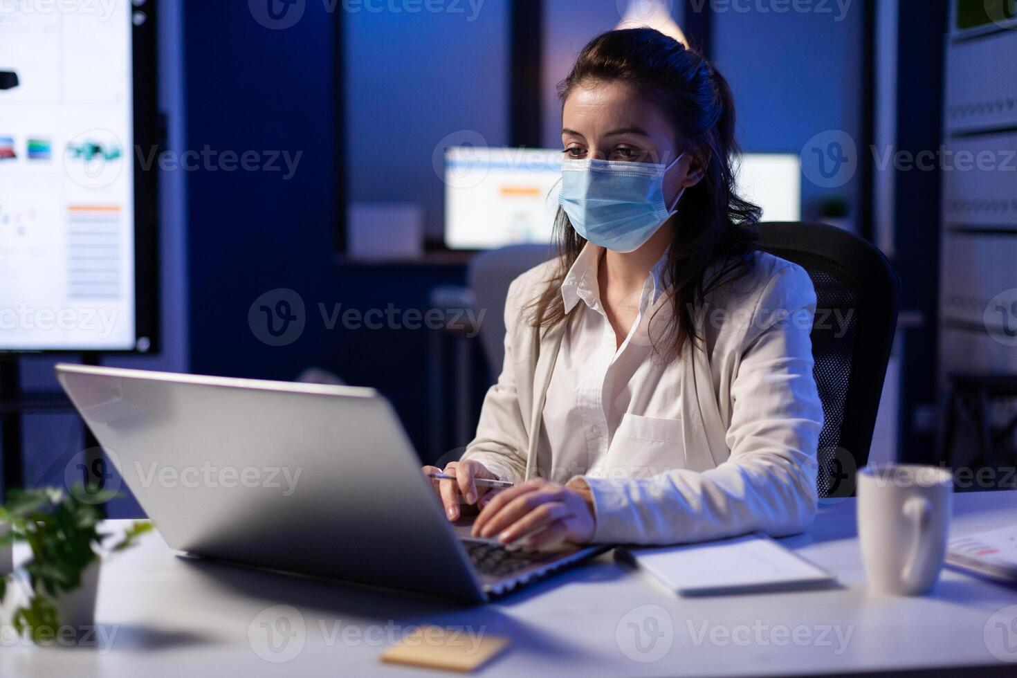 Close up of businesswoman with face mask checking emails late at night in new normal business office before deadline. Taking notes, analysing documents overtime during global pandemic photo
