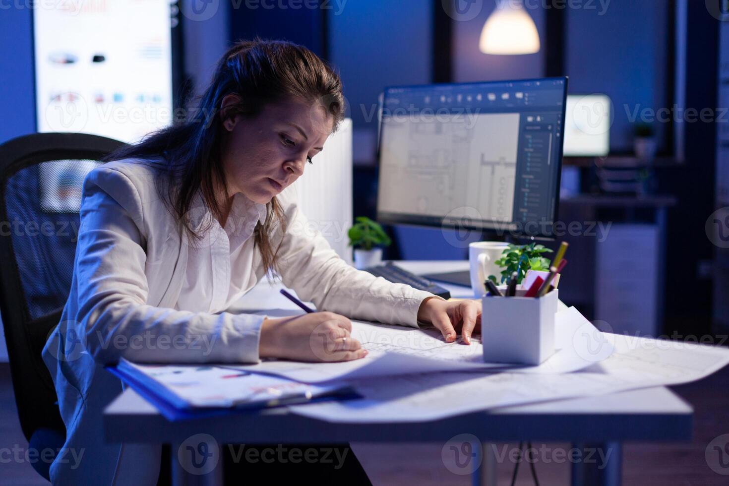 Overworked woman architect checking and matching blueprints sitting at office desk in front of computer. Perfectionist designer using arhitecture blueprints of buildings creating industrial prototype photo