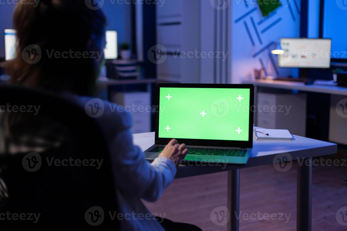 Back view of business woman looking at green screen laptop sitting at desk in start up company office . Exectutive manager watching desktop monitor display with green mockup, chroma key photo