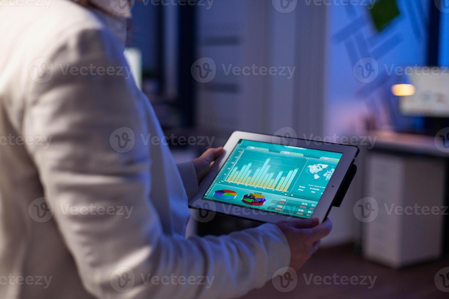 Close-up of woman hands typing on tablet checking financial graphs standing in start-up office late at night. Manager using social network, texting and blogging working overtime for job photo