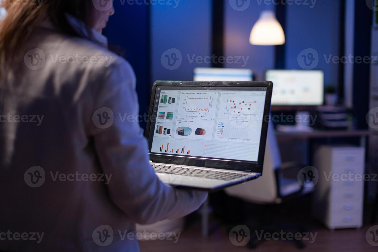 Back view of manager woman analysing financial statistics holding laptop standing in start-up business office late at night overworking. Tired focused employee using modern technology network wireless photo