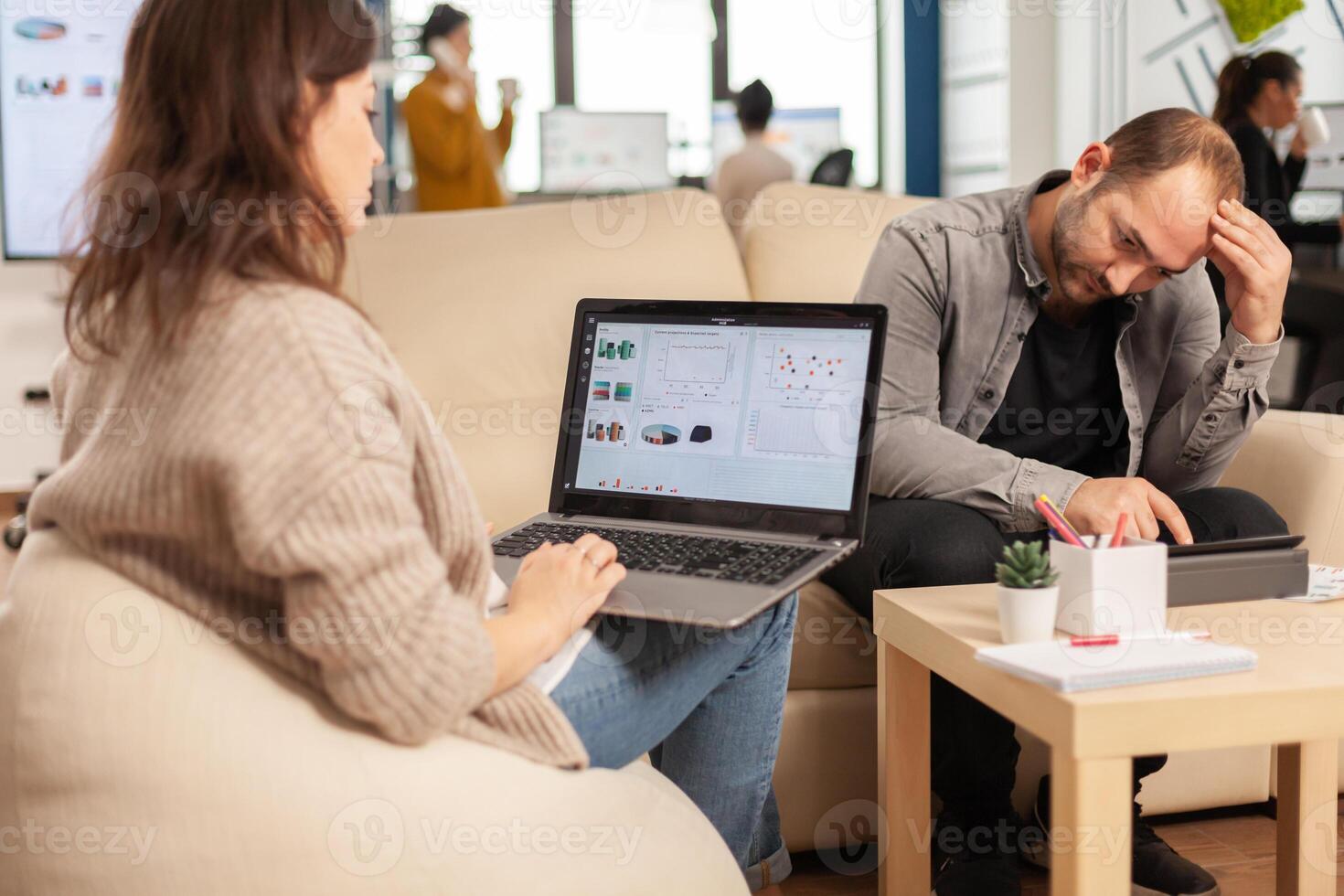 Back view of young business woman sitting on couch in office using laptop computer with graphics while colleagues working on background. Multiethnic coworkers planning new financial project photo