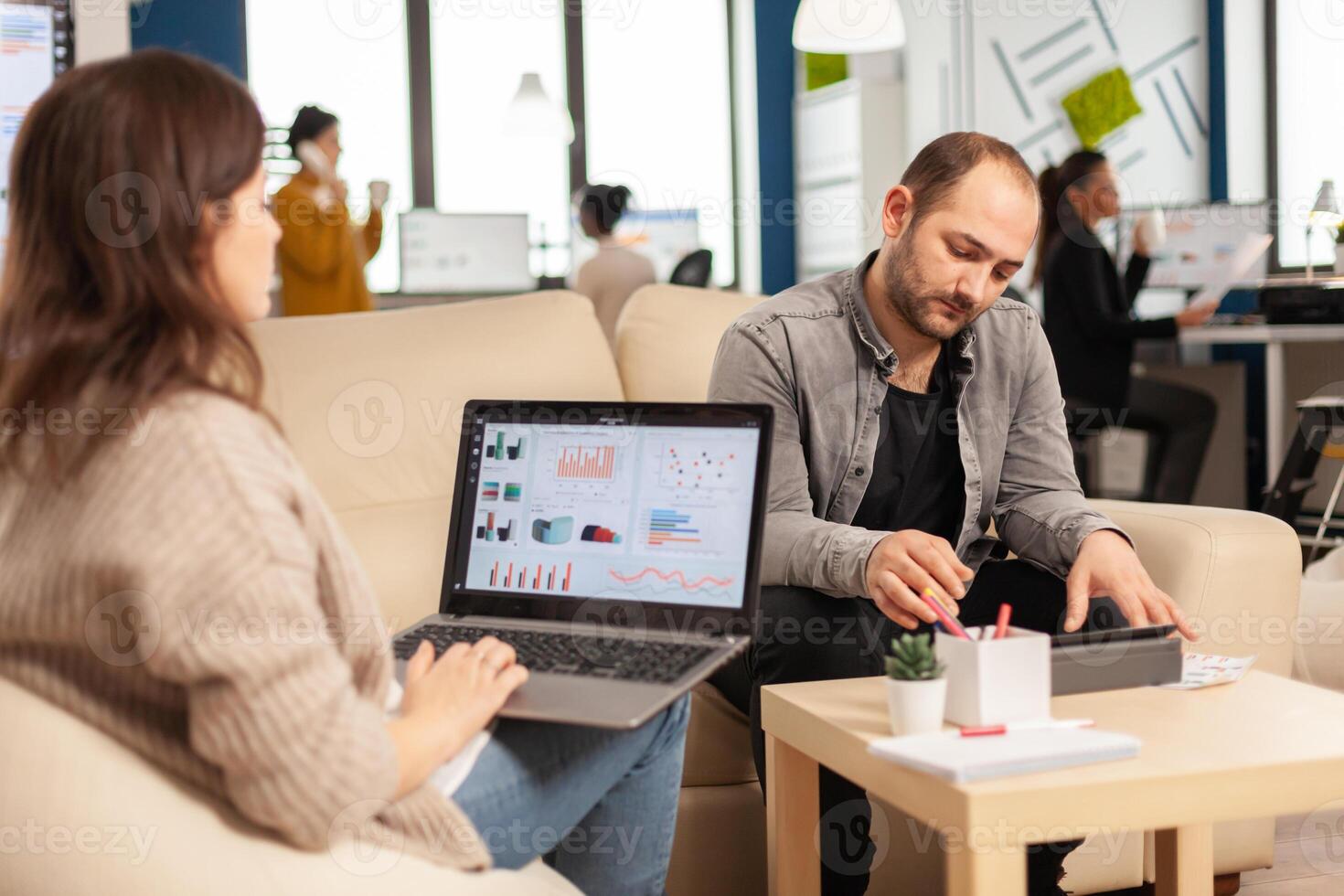 Back view of woman entrepreneur sitting on couch in office using laptop computer while colleagues working on background. Multiethnic coworkers planning new financial project in modern company photo