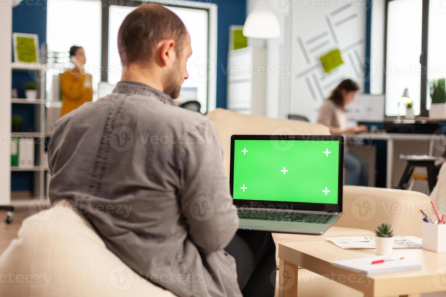 Business team sitting on couch analysing financial statistics, holding laptop with green screen while diverse team working on background. Multiethnic coworkers planning project on chroma key display photo