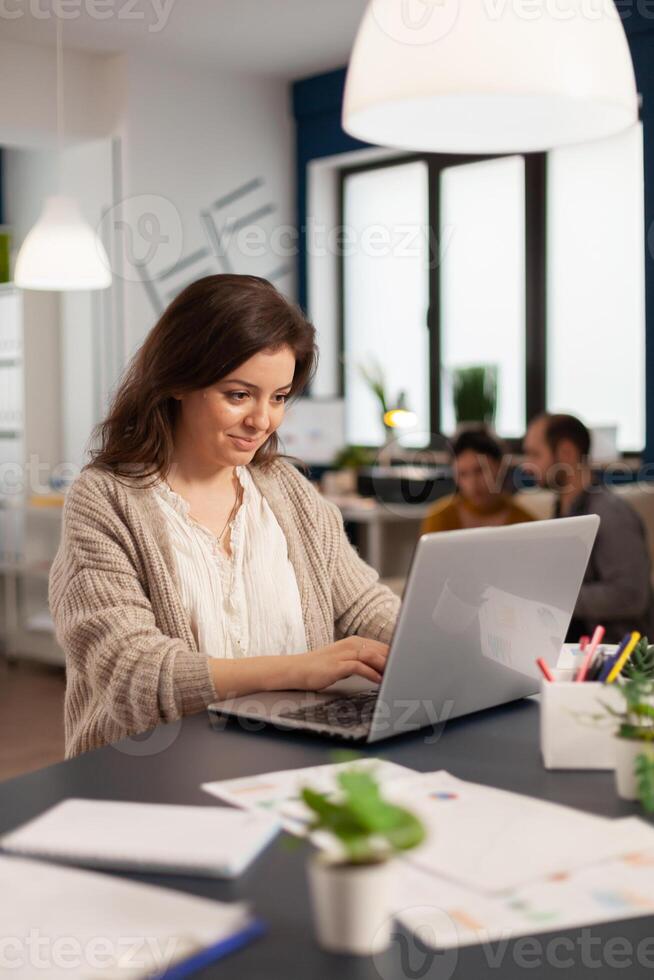 Satisfied manager checking financial statistics and graphs typing on laptop sitting at desk working in business start up company. Diverse team analyzes statistics data in modern office photo