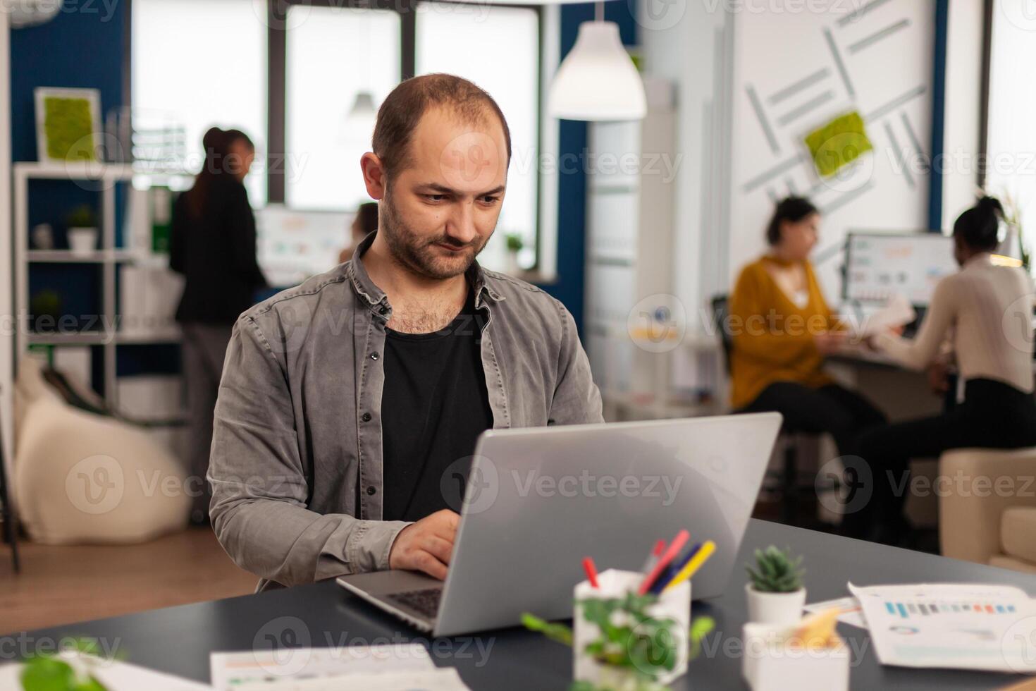 Man authentic manager sitting at table in modern startup business office sending mails typing on laptop. Business employee working in financial company with diverse team photo