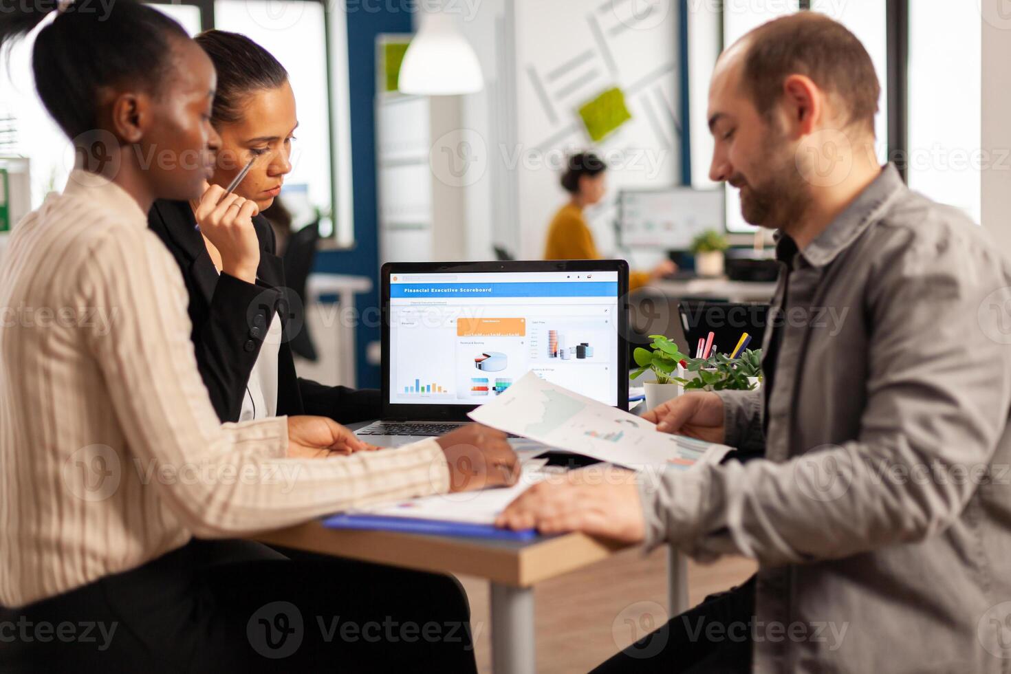 Busy multicultural, diverse employees analysing annual financial statistics sitting at desk in front of laptop holding documents searching business solutions. Team of businesspeople working in company photo