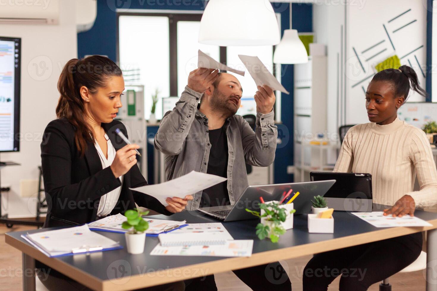 Angry business man screaming at diverse colleagues during office meeting throwing away documents, disagreeing about bad business contract. Young multiracial group of people working in coworking space. photo