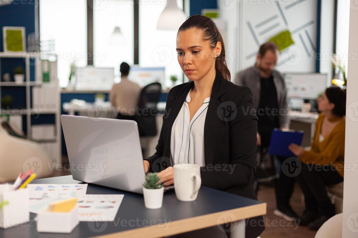 Concentrated business lady writing on laptop computer financial reports sitting at desk in busy start up office enjoying work in creative workplace. Diverse team analyzes statistics data in company photo