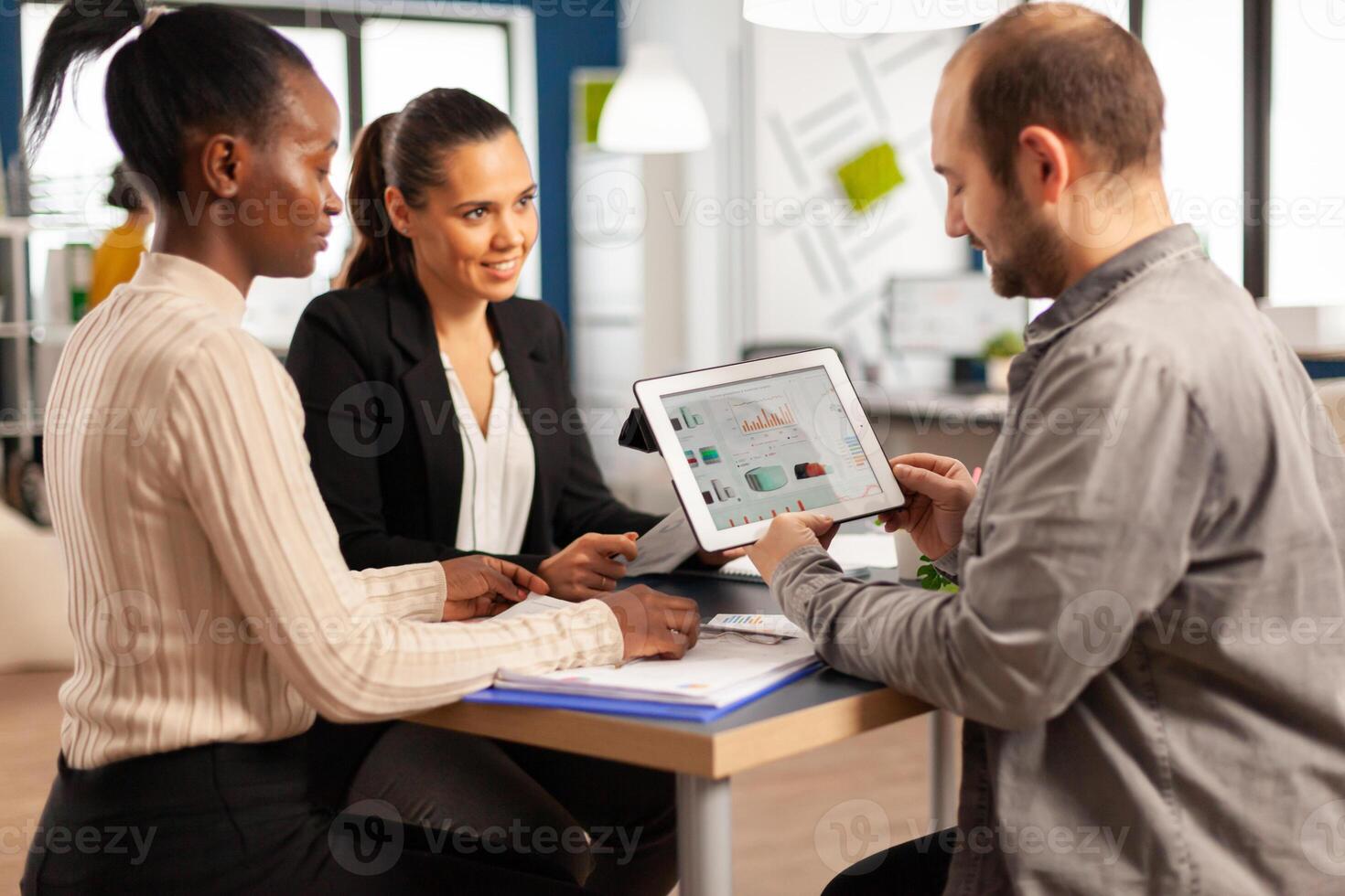 Enthusiastic diverse businesspeople reading annual financial report sitting at table in modern startup business office holding tablet and smiling. Team of multiethnic businesspeople working in company photo
