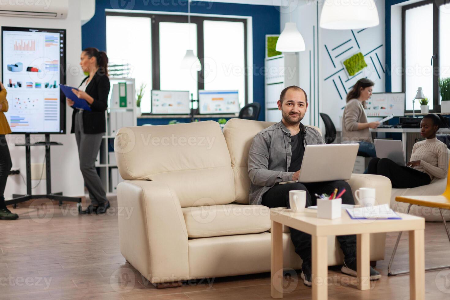 Portrait of man employee typing on laptop looking at camera smiling while diverse team working in background. Multiethnic coworkers talking about startup financial company in modern business office photo