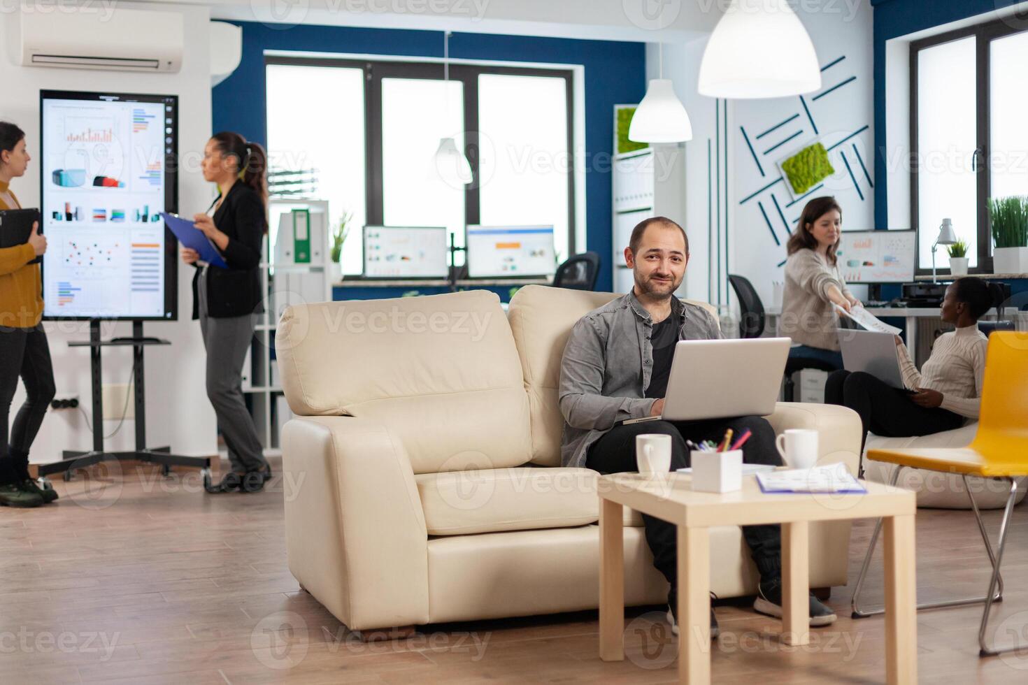 Portrait of man employee typing on laptop looking at camera smiling while diverse team working in background. Multiethnic coworkers talking about startup financial company in modern business office photo