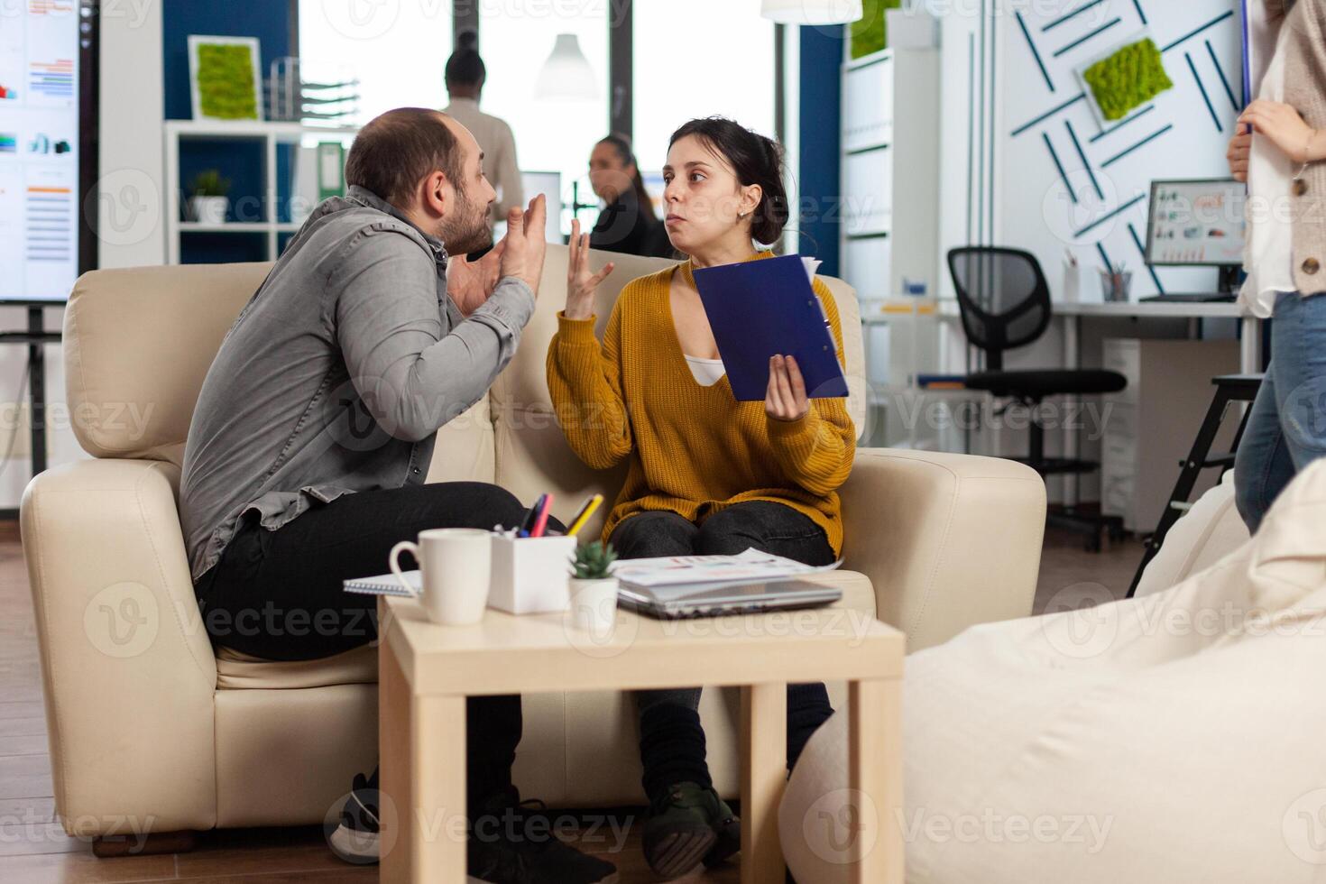 Business colleagues arguing, yealing each other during work hours sitting on the couch, while diverse colleagues working scared in background. Coworkers having conflict because of mistakes from project photo