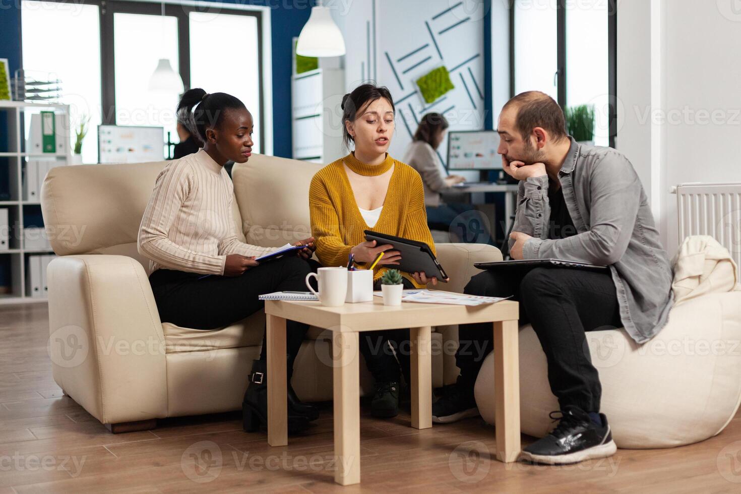 Concentrated multiethnic businesspeople reading annual financial report from tablet sitting on couch in modern startup business office. Diverse team of multiethnic businesspeople working in company photo