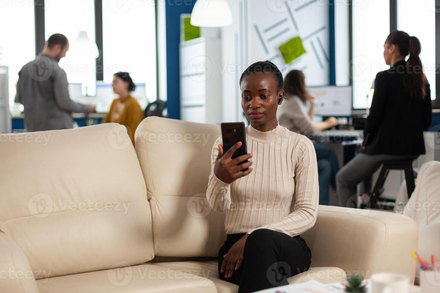 African manager woman discussing with remote colleagues on call holding smartphone using headphones sitting on couch in business modern office. Diverse coworkers planning new financial project photo