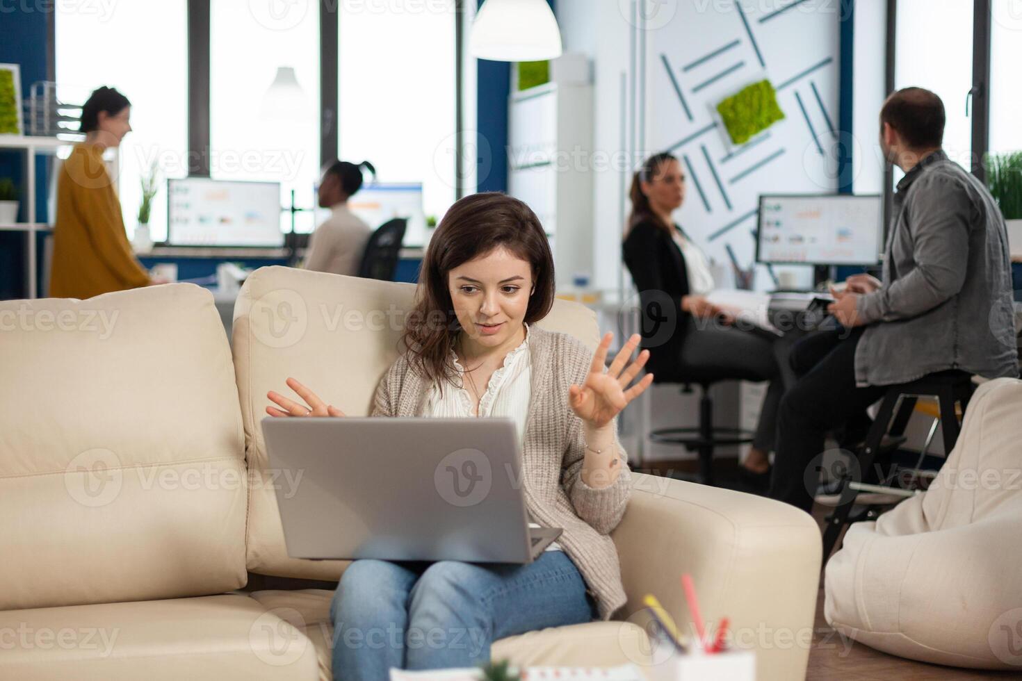 Manager woman sitting on couch holding laptop and talking on call during virtual conference working in business modern office. Diverse coworkers planning new financial project in modern company photo