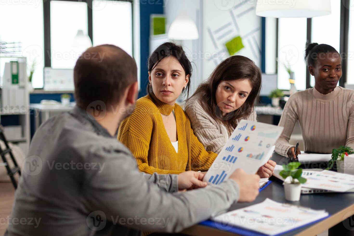 Busy multicultural women employees analysing annual financial statistics sitting at desk holding documents searching business solutions. Team of businesspeople working in company, brainstorming photo