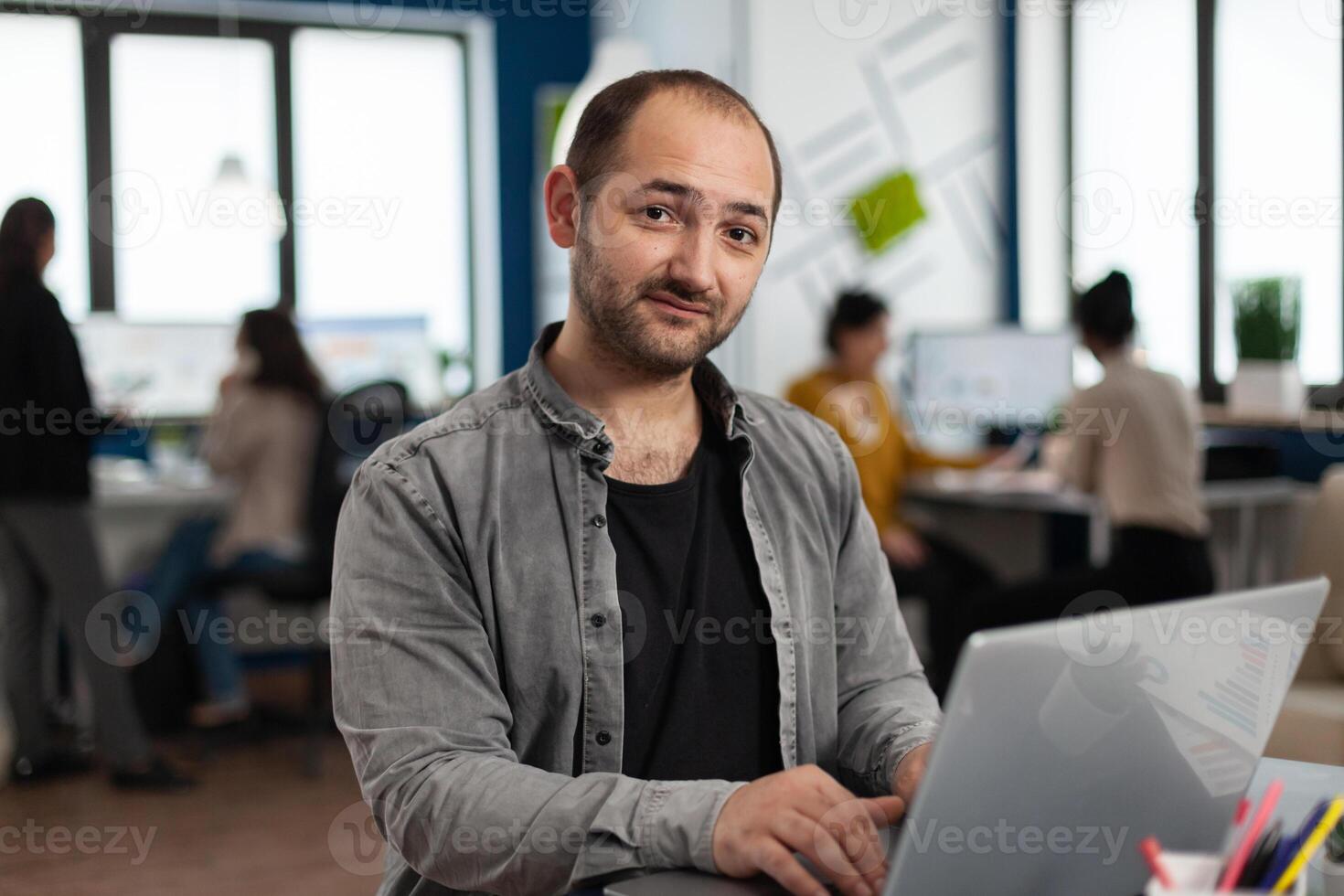 Enthusiastic business man writing a report sitting at table in modern startup business office sending mails typing on laptop, greeting colleague. Man working in financial company with diverse team. photo