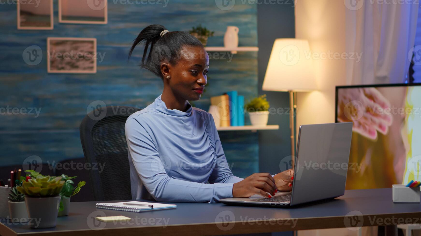 Black woman typing on computer smiling and writing informations of company on notebook sitting on desk in living room workplace late at night. African freelancer working from home overtime at laptop. photo