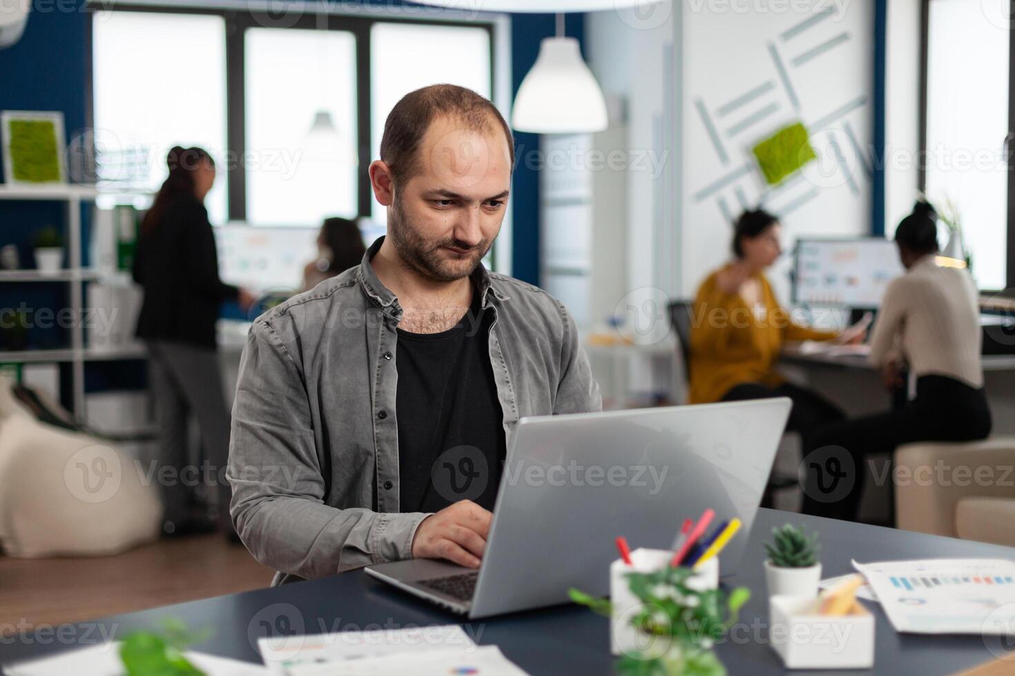 Man authentic manager sitting at table in modern startup business office sending mails typing on laptop. Business employee working in financial company with diverse team photo