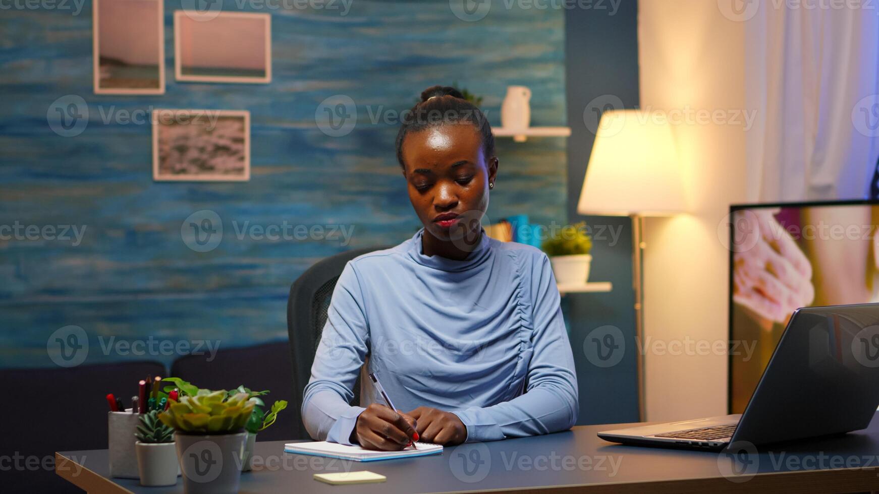 Black young business woman sitting at desk in living room workplace overtime and taking notes in notebook. African freelancer working from home late at night respecting deadline of project. photo