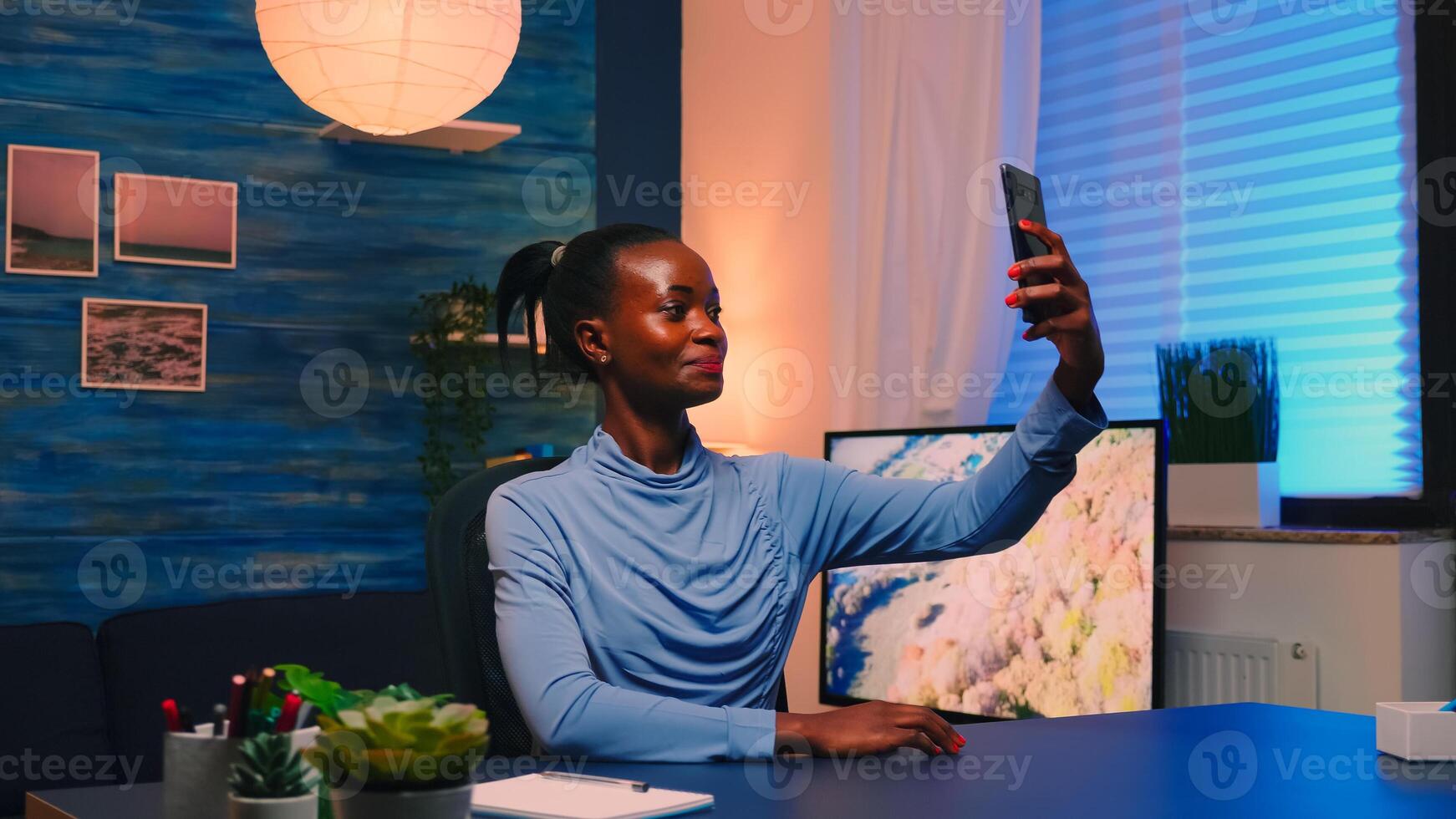African employee taking a selfie using mobile phone smiling at camera sitting at desk in living room late at night. Black woman relaxing using modern technology network wireless doing overtime for job photo