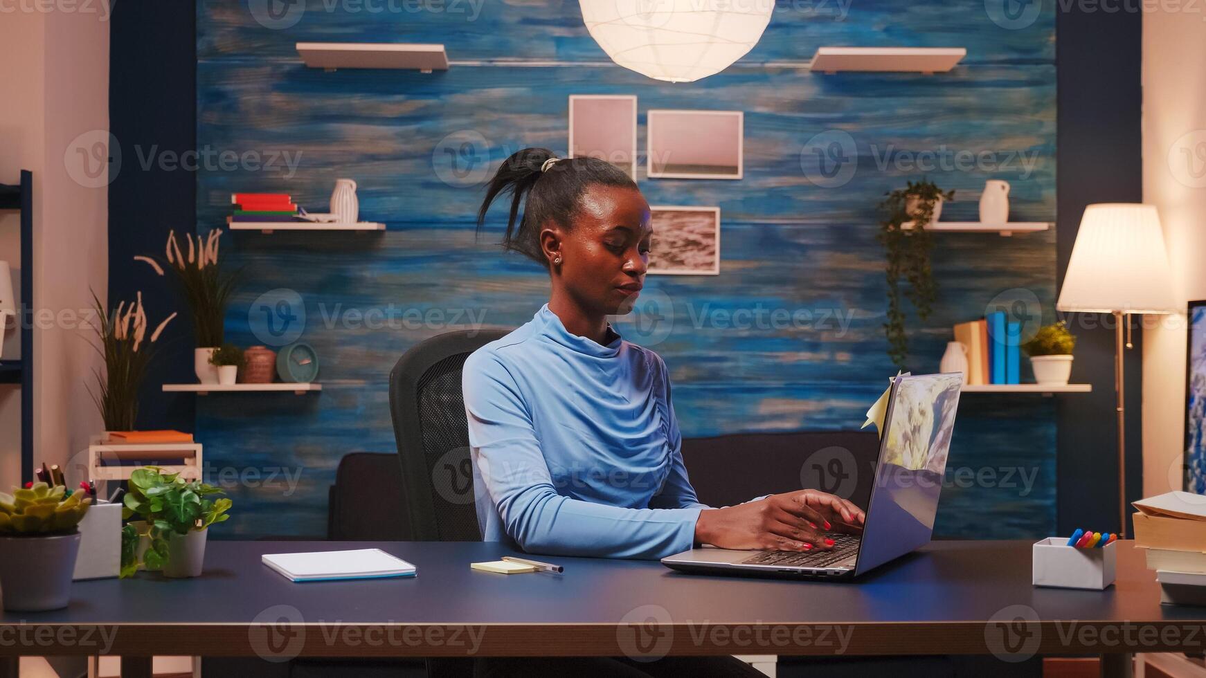Focused african freelance writing on computer and using sticky notes for do list putting it on laptop sitting on desk in living room workplace late at night. Black woman working from home overtime photo