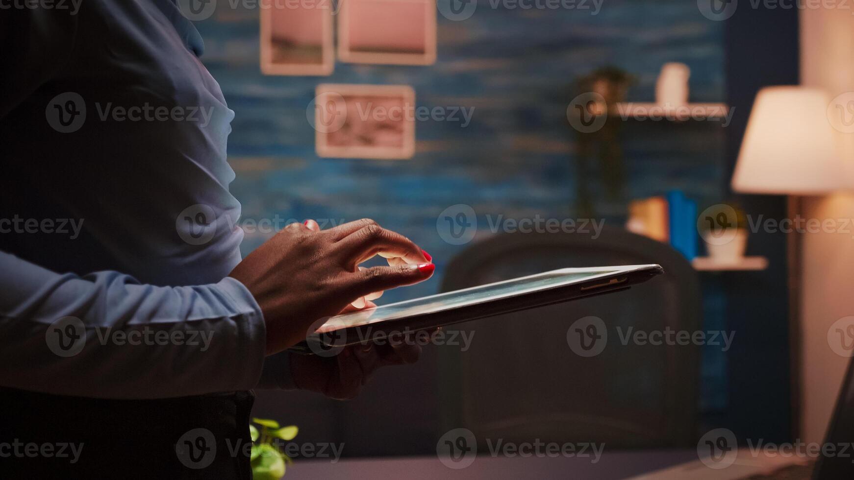 Close up of african employee texting, sending and reading messages during her break standing in living room late at night. Focused black employee using modern technology network wireless overworking photo