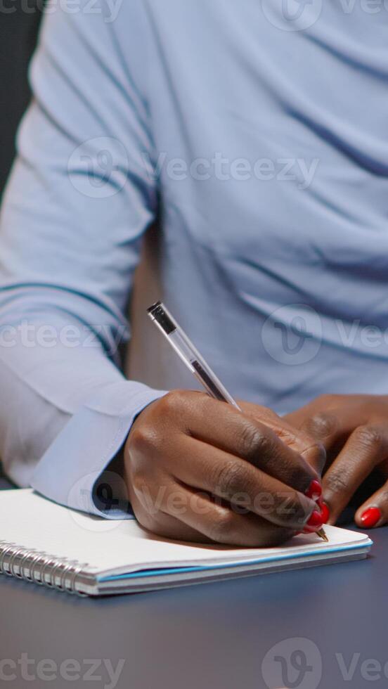 Close up of african businesswoman writing list of tasks for business project in notebook sitting at desk in living room working overtime. Black freelancer respecting deadline studying late at night photo