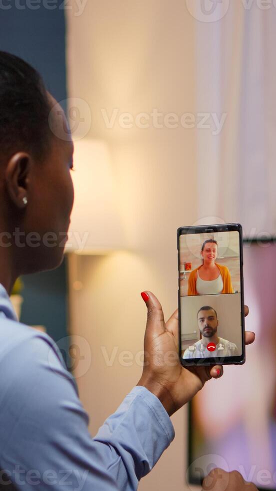 Black woman remote entrepreneur having conference with friend using smartphone sitting in living room late at night. Busy employee using modern technology network wireless doing overtime for job photo