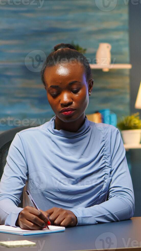 Black young business woman sitting at desk in living room workplace overtime and taking notes in notebook. African freelancer working from home late at night respecting deadline of project. photo