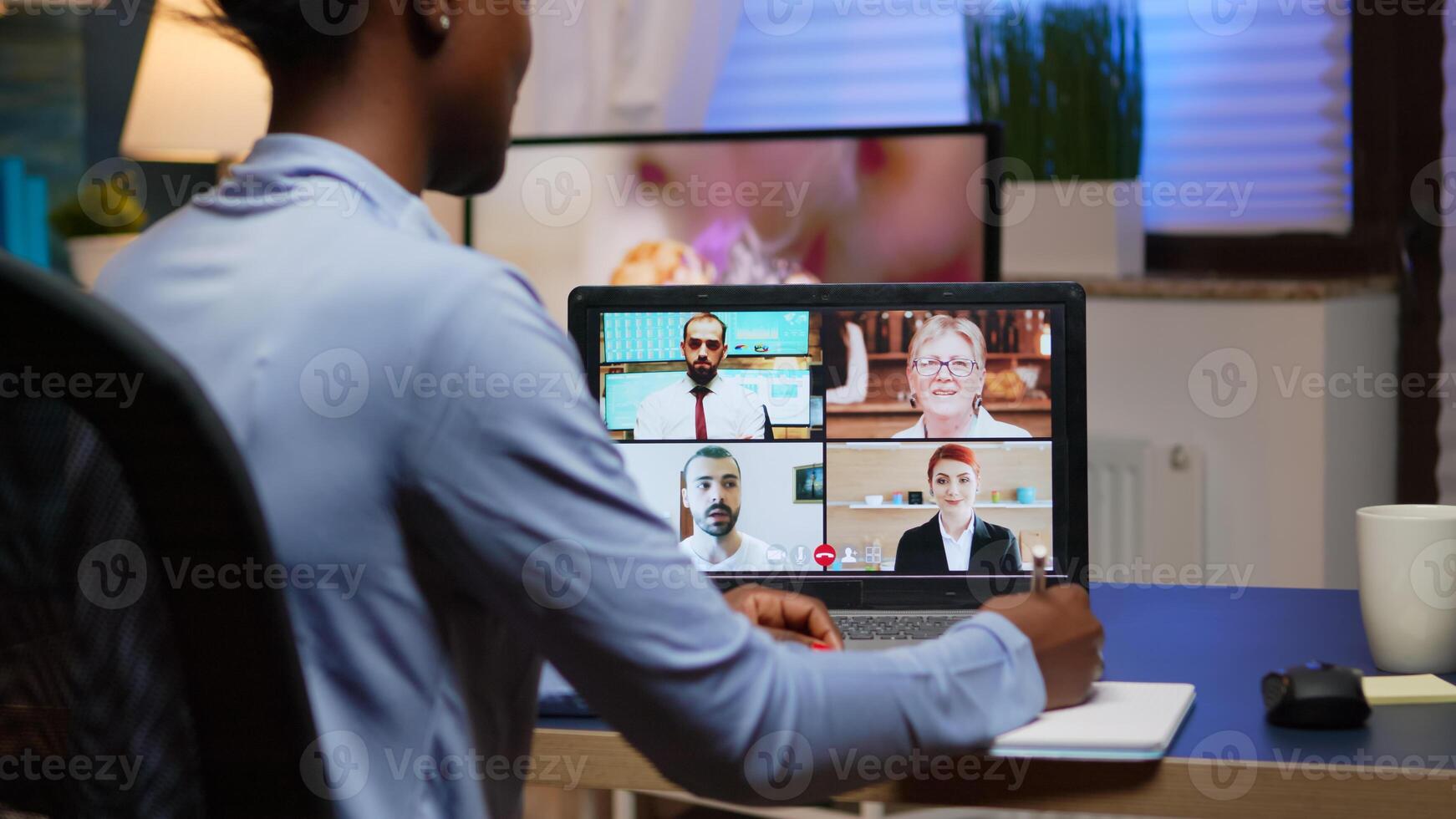 African freelancer working remotely discussing with partners online using laptop sitting in Livin room. Using modern technology network wireless talking on virtual meeting at midnight doing overtime photo