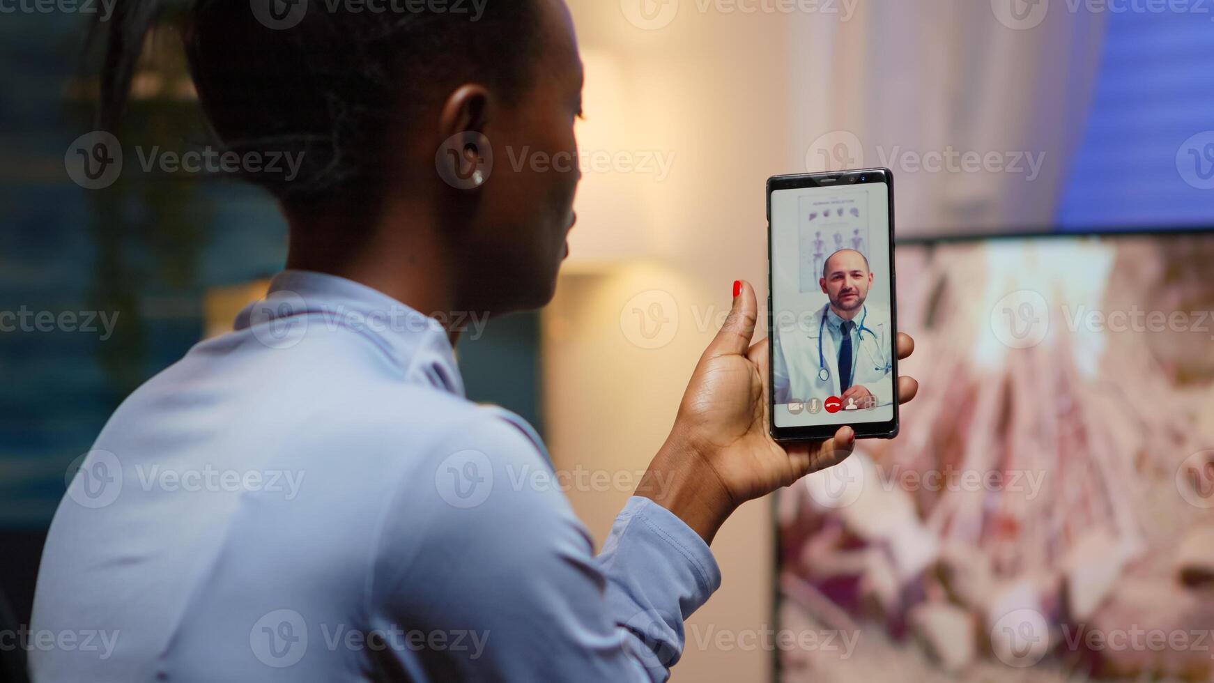 African famele having conference call with her doctor about illness treatment sitting in living room late at night. Black patient in a call with medic discussing health problems of woman photo