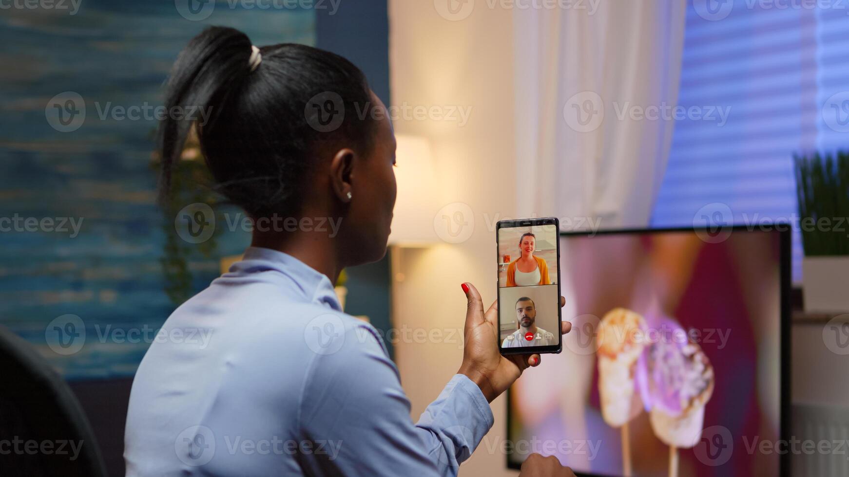 Black woman remote entrepreneur having conference with friend using smartphone sitting in living room late at night. Busy employee using modern technology network wireless doing overtime for job photo