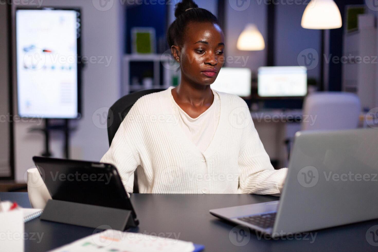 Workaholic african manager trying to finish deadline checking charts on tablet pc and laptop. Busy multitasking employee analysing financial statistics overworking writing, searching. photo