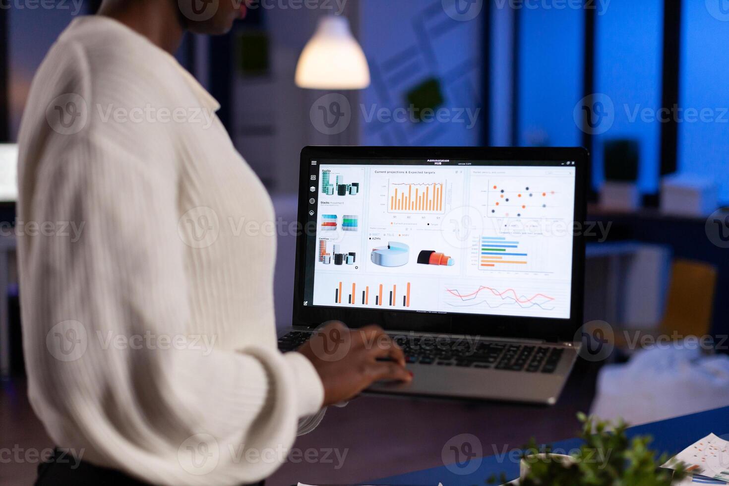 Back view of african business woman analysing financial statistics holding laptop late at night. Busy focused employee using modern technology network wireless overtime. photo