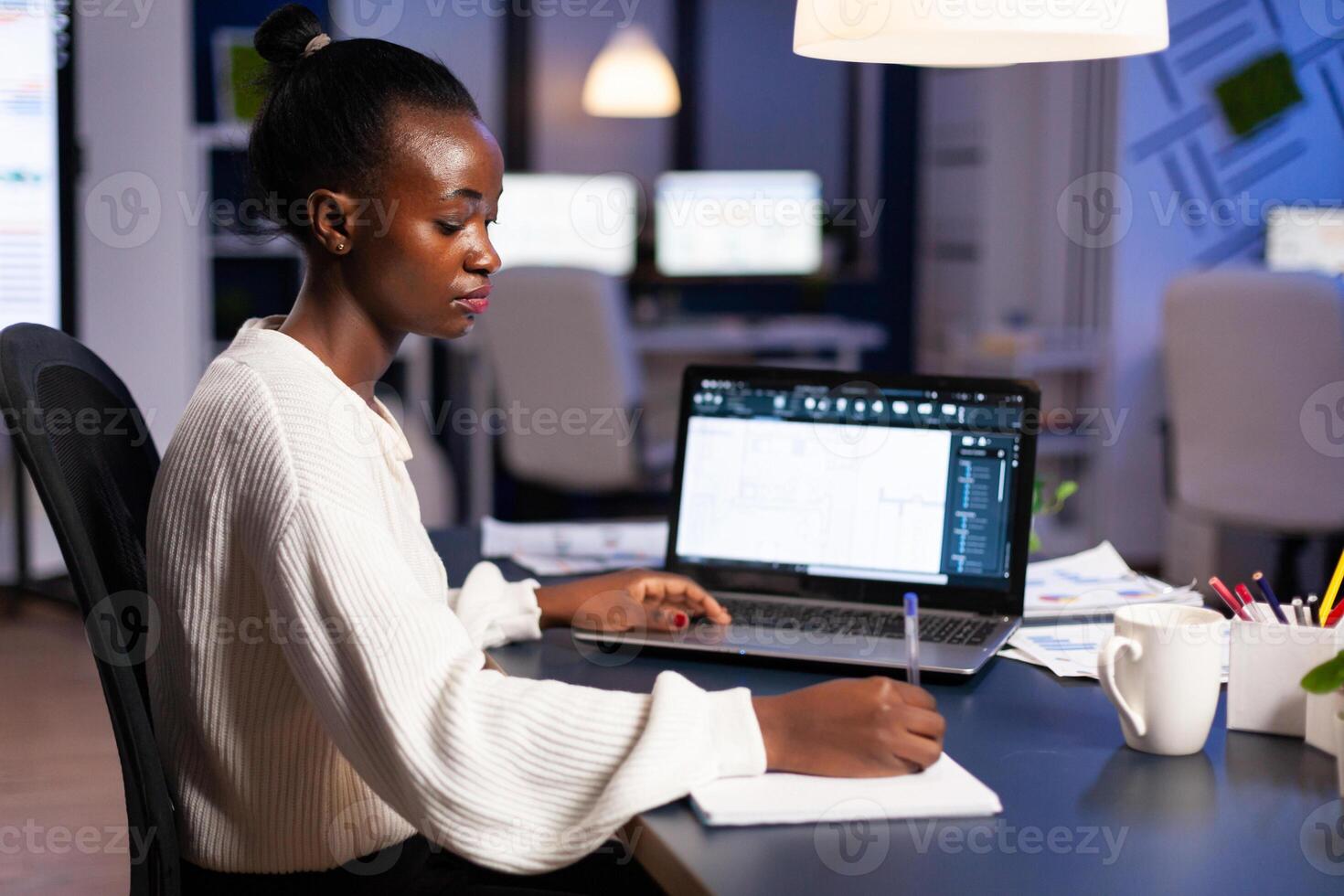African architect taking notest on notebook working on project looking at blue prints. Industrial woman employee studying prototype idea showing cad software on device display. photo