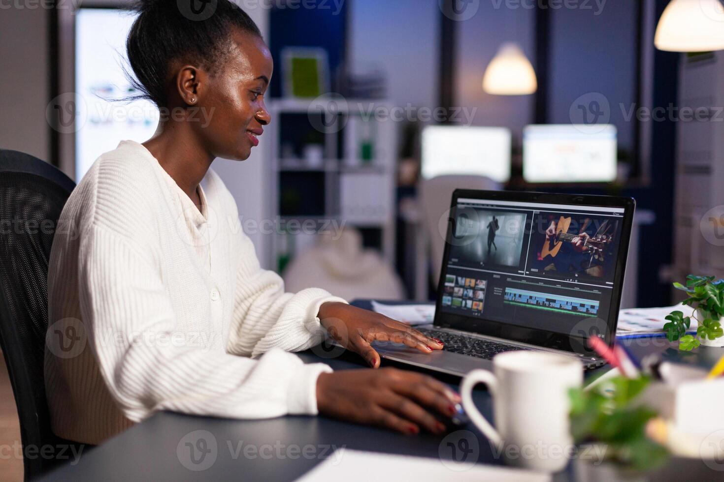 Black delighted videographer editing movie on professional laptop sitting at desk in business office at midnight. Creative editor working at night at new project processing audio film montage. photo
