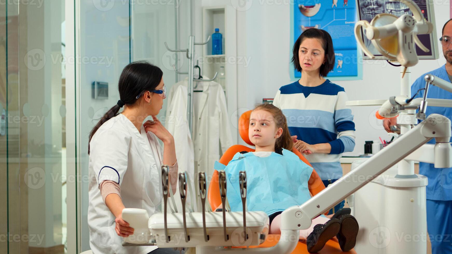 Stomatologist doctor explaining to mother the dental process of intervention for teeth problems of kid, girl showing to affected mass. Orthodontist speaking to child sitting on stomatological chair photo