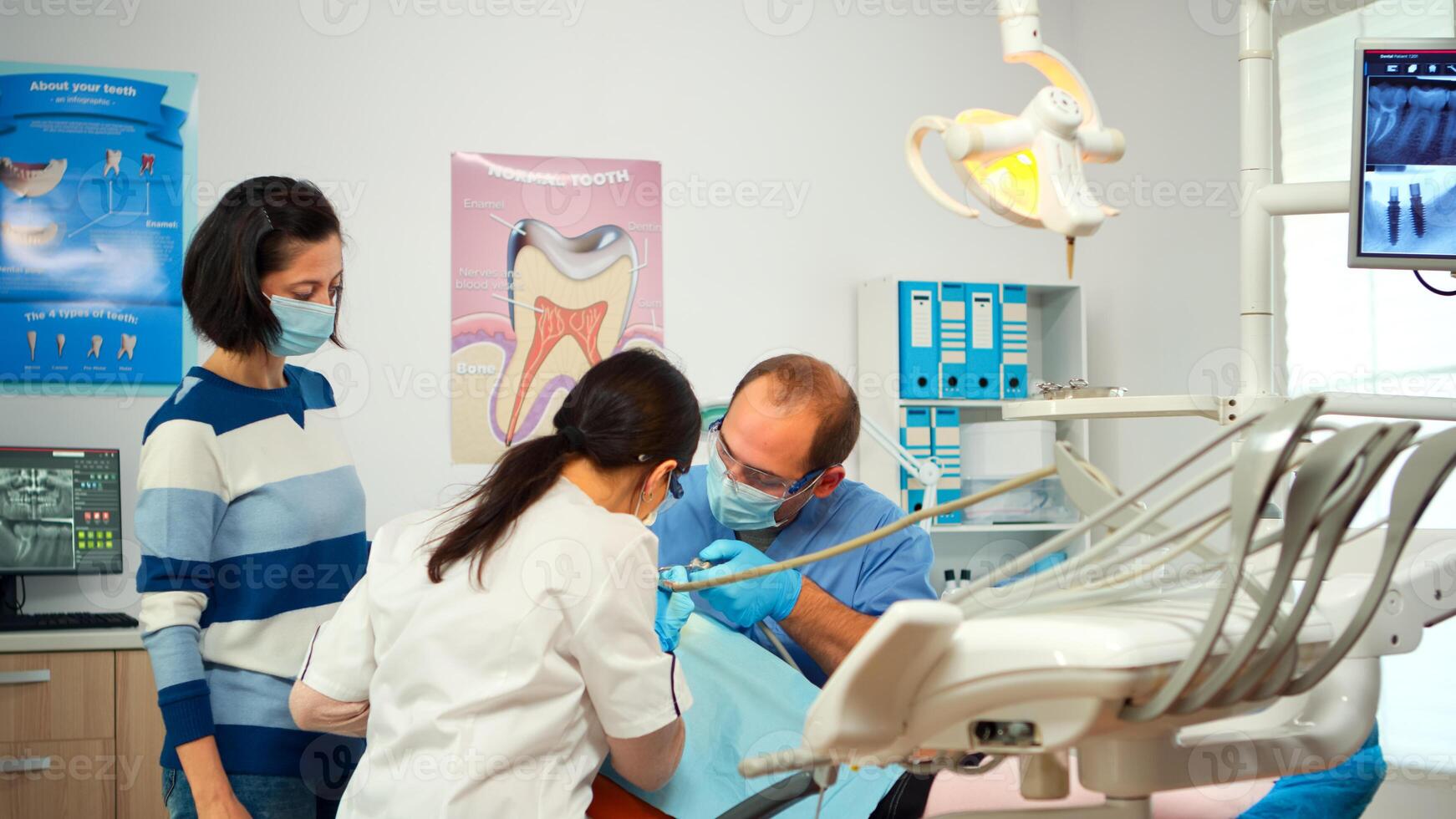 Little girl patient sitting in stomatological chair in dental office, visiting doctor for oral problems. Woman dentist with protective gloves and mask removes tooth decay drilling teeth during surgery photo