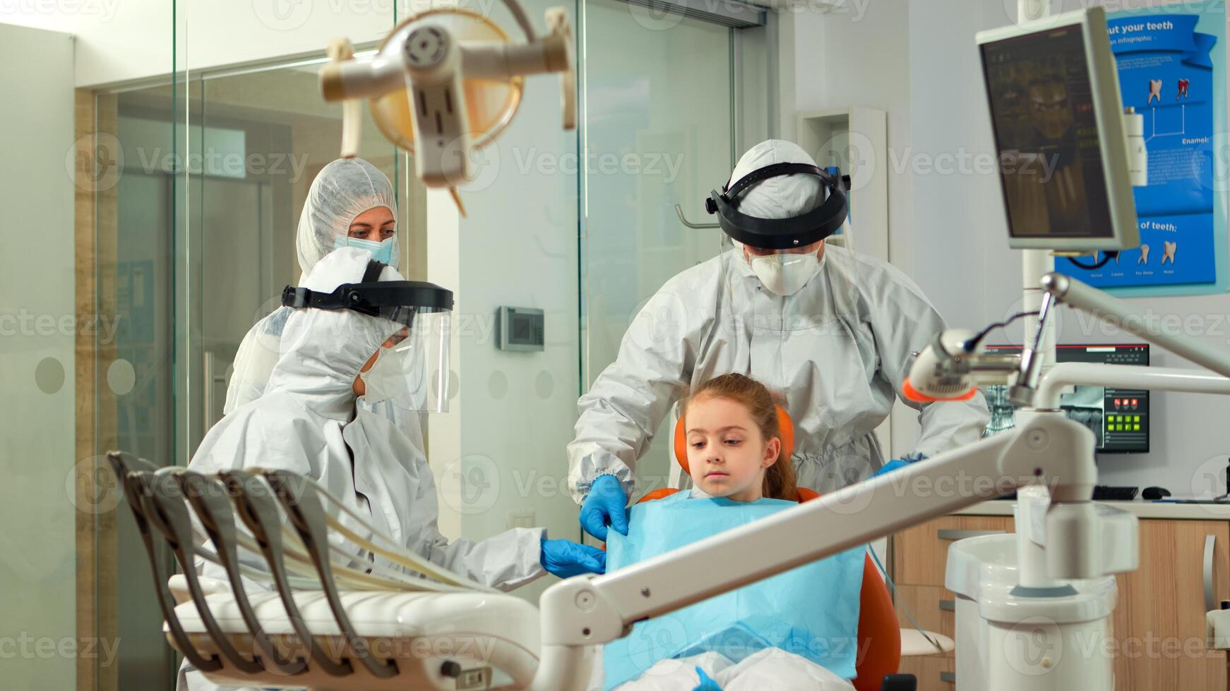Dentistry nurse in coverall putting dental bib to child before stomatological examination during covid-19 pandemic. Concept of new normal dentist visit in coronavirus outbreak wearing protective suit photo